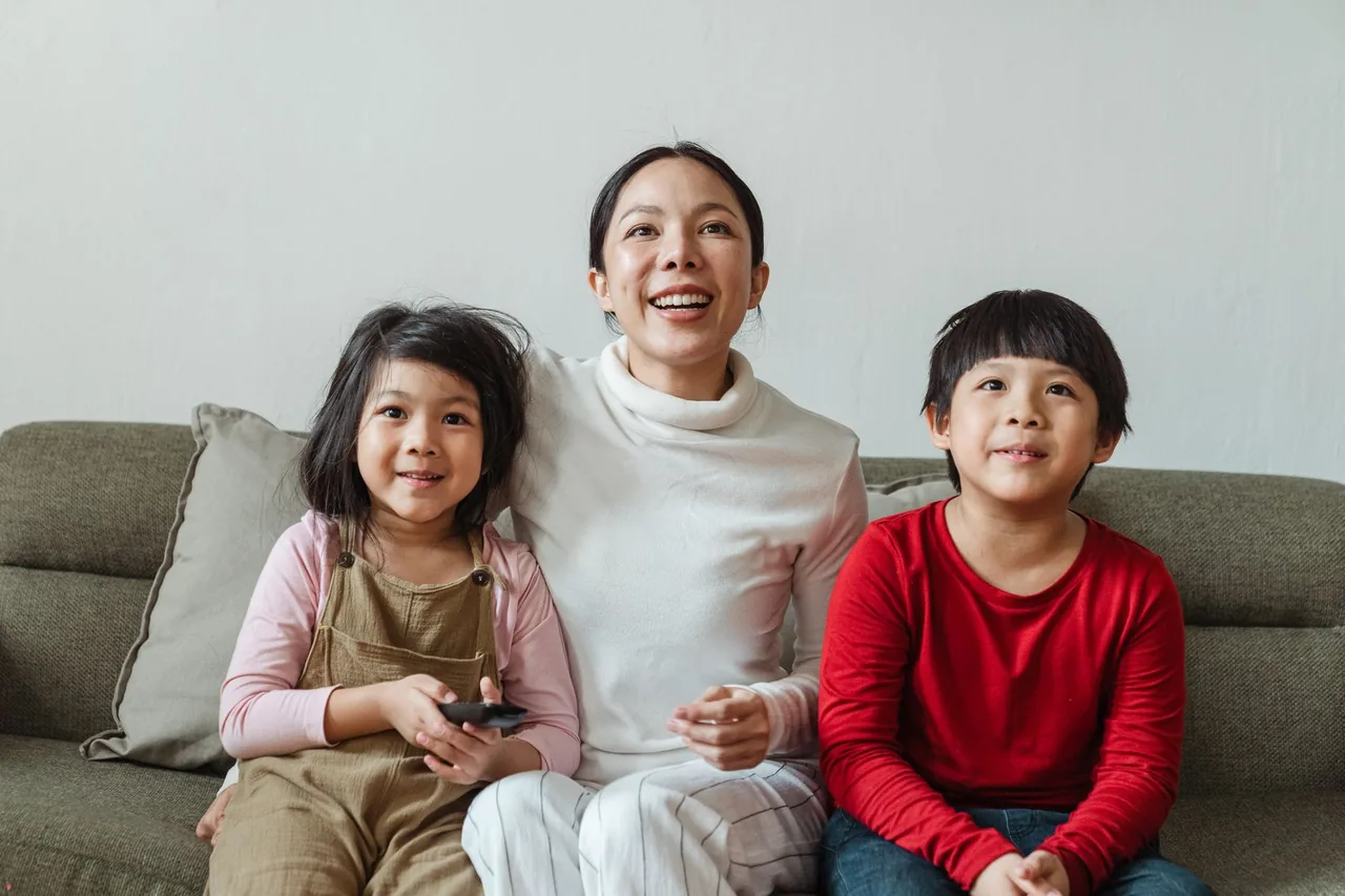 A laughing woman on a sofa between two children, the girl holding the TV remote