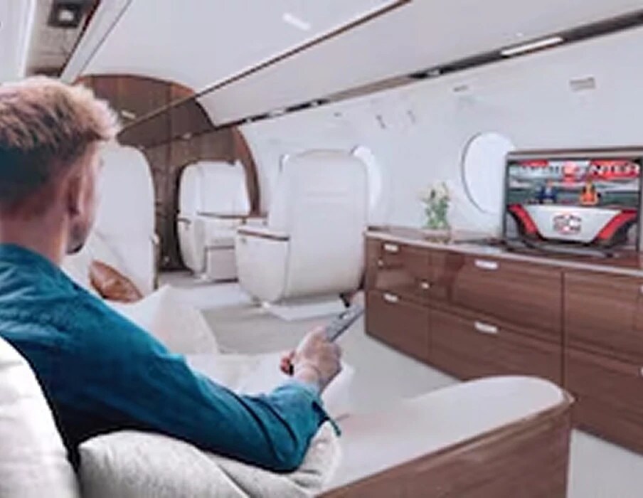 A man relaxing in a white leather seat of a private jet, pointing a remote at a TV across the cabin
