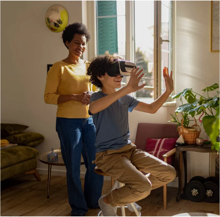 A woman in a yellow sweater laughing as a boy on a stool tries out a VR headset in a sunny living room